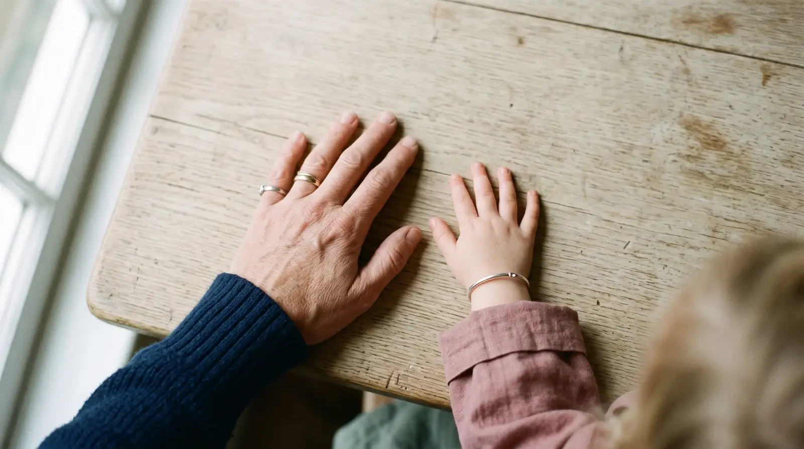 Mano de una persona adulta y mano de un niño apoyadas juntas sobre una mesa de madera clara